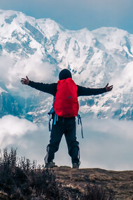 A man stands with both hands raised on a mountain cliff, overlooking the snow-covered peaks of the Annapurna range in Nepal