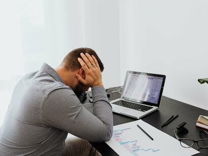 Sentado diante do computador portátil e de documentos com gráficos, o homem segura a cabeça com as mãos, num gesto de preocupação. A cena remete às consequências de escolhas que comprometem o futuro financeiro e profissional, tema presente na crítica do autor às promessas ilusórias e aos riscos de acreditar em discursos sem fundamento