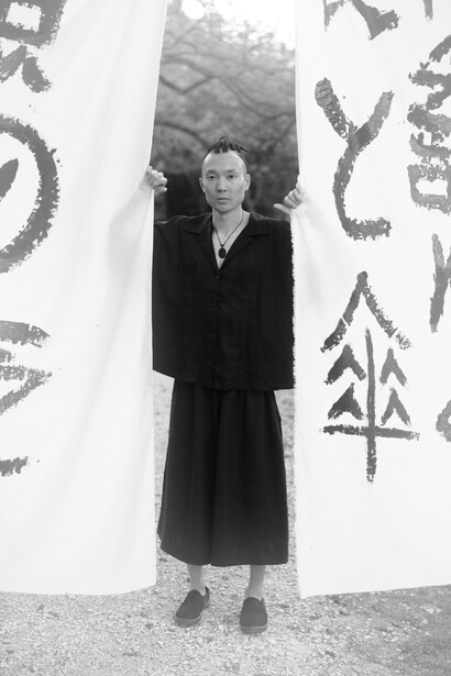 Hombre japonés vestido de negro parado detrás de telas con una escritura pintada