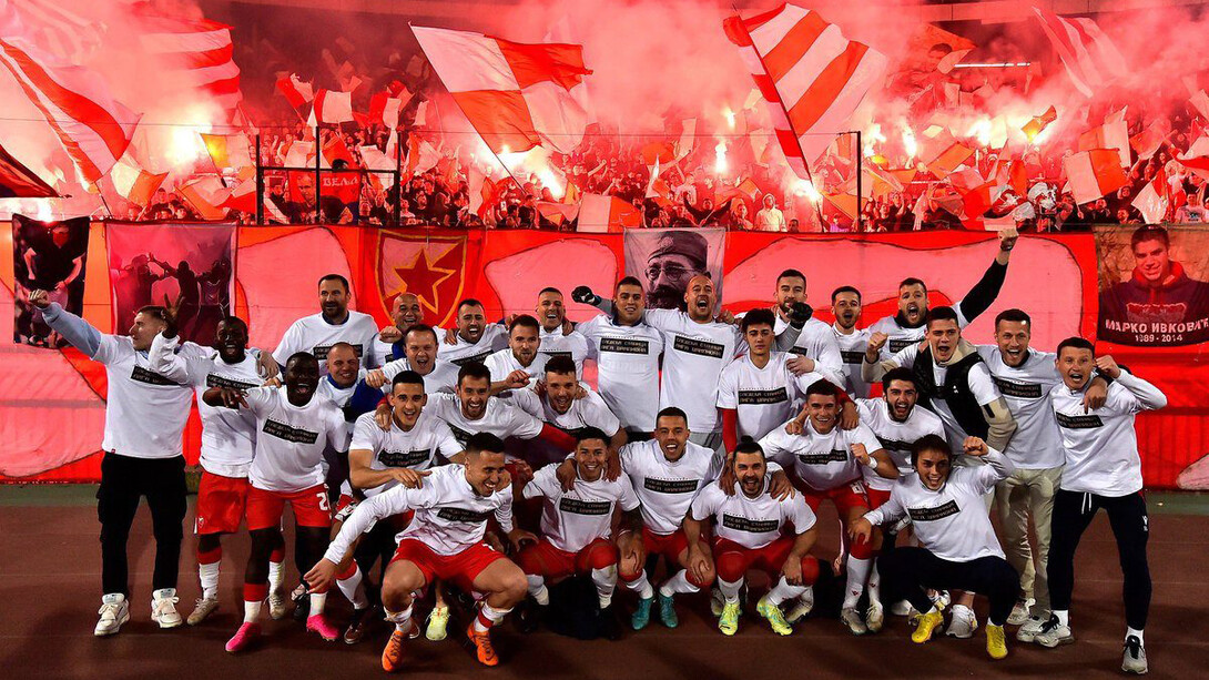 Jogadores do time Estre Vermelha, seleção de fuebol da República da Sérvia, juntos em foto enquanto a torcida ultra ao fundo comemora com a tradicicional queima de artifícios pirotécnicos