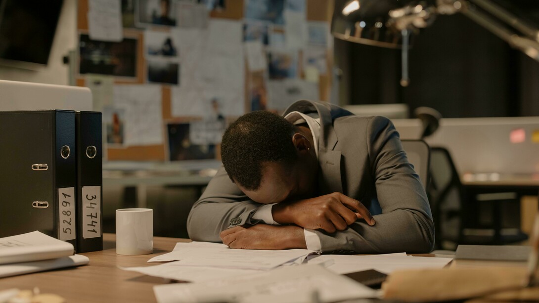 A man in a gray long-sleeved shirt rests his head on the table, crying at work
