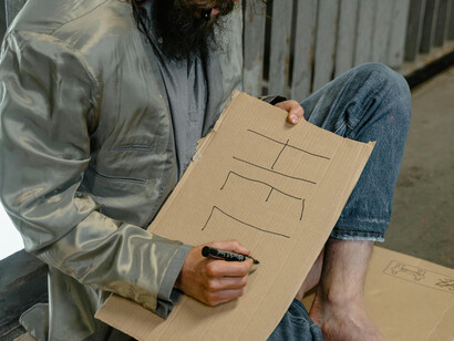Seated on the sidewalk, a male beggar holds a banner requesting help from passersby