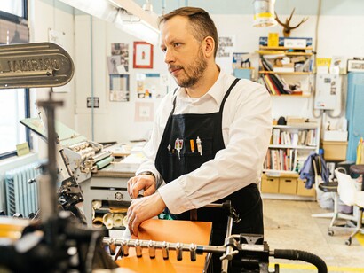 Homem trabalha diante de uma prensa de impressão, acompanhando o equipamento durante o processo de produção. A imagem remete à experiência do autor nas oficinas gráficas, onde o conhecimento técnico e a atenção aos detalhes eram fundamentais para o trabalho