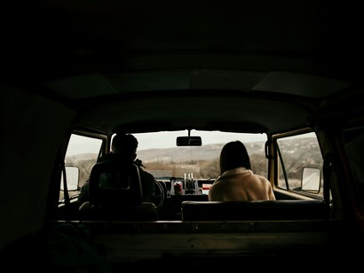 Seen from the back of a van, two travelers sit in silence as the road stretches ahead, turning movement into a shared, reflective experience rather than just transit