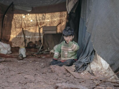 Inside a fragile tent, a child sits amid mud and debris—an image that reflects the broken promises of development programs that fail to reach those in need