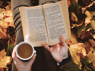 A person holds a cup of coffee while reading a book among autumn leaves