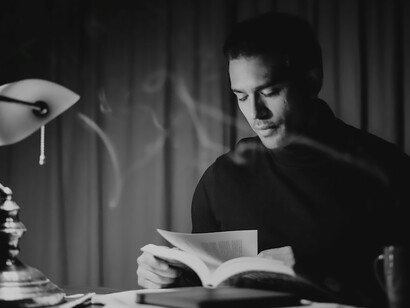 Dressed in a black sweater, a contemplative man sits in a black and white room, lost in profound thought while earnestly crafting the pages of a book