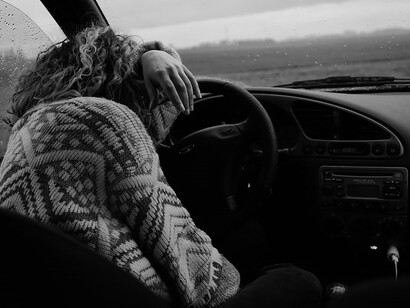 An individual, donning a somber gray and white sweater, drives a car with a sense of despondency coloring their demeanor