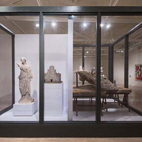 It Takes a Village, (2015), is a new, site-specific work installed in a large glass vitrine and includes various found objects, including Mexican clay figurines, artificial aloes, a plastic radio, a marble statue, Spanish moss, and Mexican colonial painting, among others