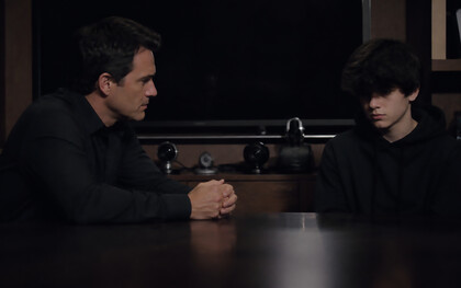 A father and son sit across from each other in silence, their tension shaped by control, performance, and the unseen weight of being watched