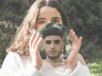 Amidst the lush greenery, a couple stands, and in the girl's hand, a mirror reflects the image of the man