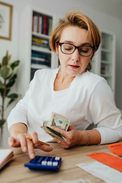 A woman counting money and receipts with a calculator at a kitchen table