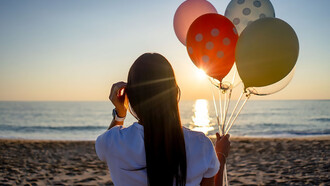 A woman standing on the beach, holding colorful balloons, with her back to the camera