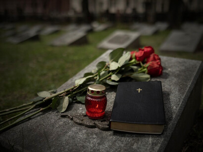 A gravestone adorned with flowers stands in a cemetery as a burial ceremony takes place, with mourners gathered and a funeral procession in the background