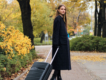 Side view of a thoughtfully dressed young woman walking through a city street with a small black suitcase, symbolizing emigration and the modern nomadic lifestyle