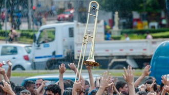 Aglomeração de pessoas com um trombone em destaque entre a multidão. A imagem remete à expressão “botar a boca no trombone”, usada quando alguém torna público aquilo que pensa, denuncia uma situação ou decide falar abertamente sobre algo — um exemplo de como o sentido figurado transforma uma expressão cotidiana