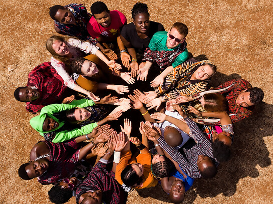People from diverse backgrounds gather in a circle in Masimba, Tanzania, expressing solidarity and community through a shared gesture of unity