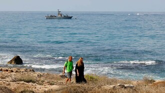 People walking along the beach as a navy vessel patrols the Mediterranean waters off Rosh Hanikra, known in Lebanon as Ras al-Naqura, on the Israeli side of the border between the two countries, on October 7, 2022