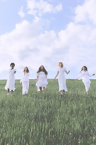 Amidst the natural expanse, five women in white dresses run hand in hand, their laughter echoing through the air, creating a timeless moment of harmony and joy