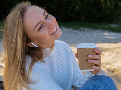 On the beach, a content woman sips coffee, headphones on, exploring self-discovery through audio healing, nature, and mindfulness