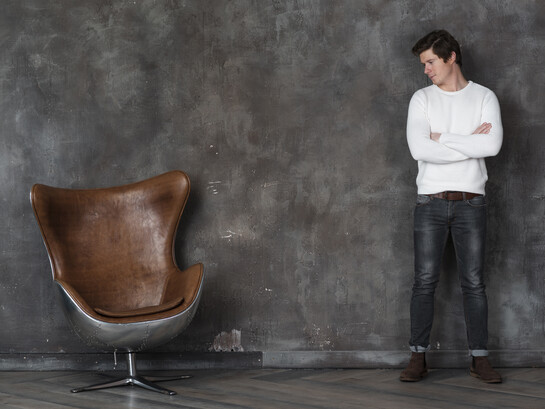 A man stands facing an empty leather armchair, appearing reflective and contemplative