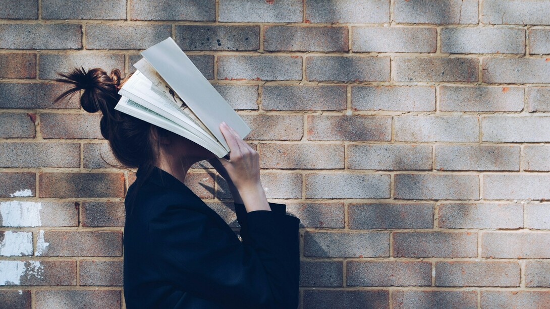 A schoolgirl stands against a rustic red brick wall, holding a book up to cover her face, reflecting student burnout