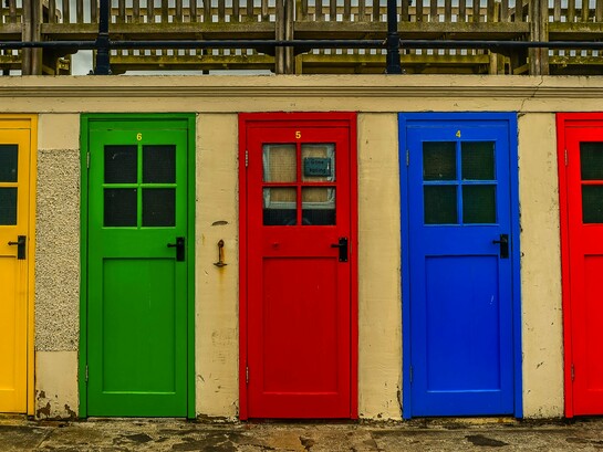 Uma sequência de portas de diferentes cores se alinha lado a lado em meio à paisagem urbana, criando uma composição marcada pela diversidade. Cada porta pode ser apenas uma escolha estética, mas também pode dizer algo sobre quem está do outro lado — afinal, como pergunta a autora, qual é a cor da porta da sua casa?