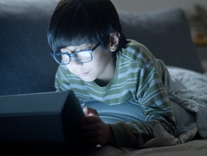 A boy in glasses concentrates on a digital tablet in a dimly lit room at night