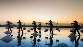 Salt harvesters carrying baskets across shallow waters at sunrise, representing the dignity and value of labor in sustaining society