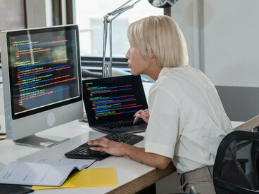 Mulher trabalha diante de múltiplas linhas de código exibidas no monitor. A construção de uma IA mais justa depende também de quem participa do seu desenvolvimento. O artigo relaciona representatividade e qualidade no desenho de sistemas de inteligência artificial. Reduzir preconceitos algorítmicos exige atenção aos dados e às equipas responsáveis pela tecnologia