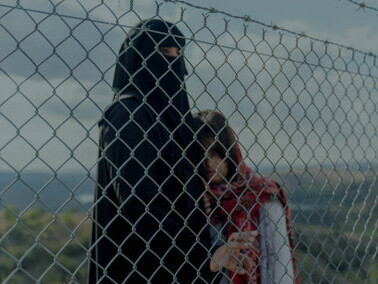 A refugee woman stands behind a fence, her gaze reflecting the uncertainty of her journey