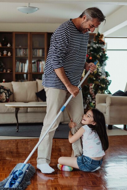 Young girl holds her fathers leg while he mops the floor