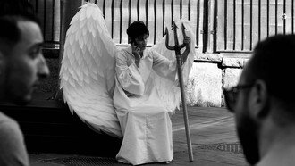 Uma pessoa caracterizada como um anjo, com grandes asas e um tridente nas mãos, sentada enquanto fala ao telefone. A imagem brinca com a dualidade entre aparência e comportamento — justamente a questão que atravessa o artigo: quem somos quando não há ninguém para nos observar? Entre aquilo que mostramos aos outros e aquilo que permanece oculto, o caráter se revela justamente quando não há ninguém para nos observar