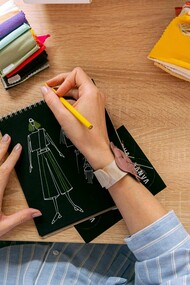 Mãos desenhando roupa em um caderno sobre uma mesa de estudos de moda. Entre traços e ideias, nasce a moda em um processo criativo que une arte, técnica e consciência em cada detalhe. O primeiro esboço de uma peça carrega mais que estética, revela pensamento, cultura e intenção. Antes das passarelas, a moda começa no papel, onde criatividade e propósito se encontram