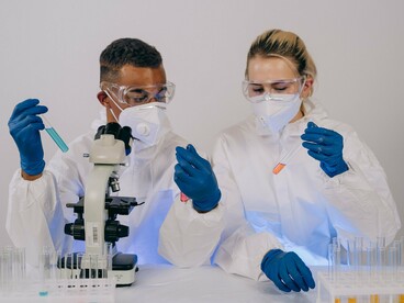 Dois profissionais realizam atividades em um laboratório, utilizando equipamentos de proteção e instrumentos de análise científica. O desenvolvimento de vacinas personalizadas de RNA mensageiro depende de pesquisas que buscam identificar características específicas de cada tumor e transformá-las em alvos para a resposta imunológica
