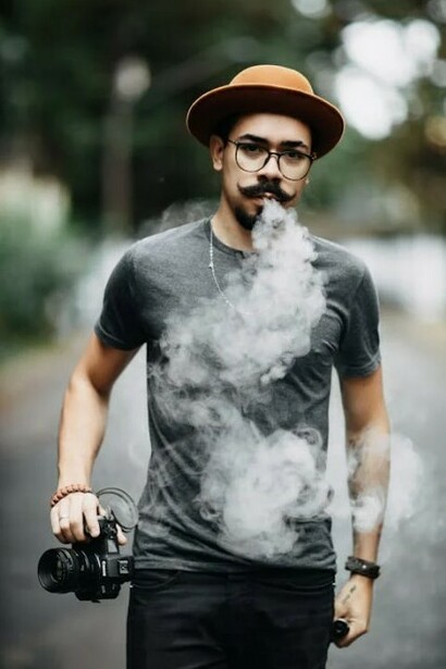 A man posing in a hat and glasses, with a mustache, wearing a simple t-shirt and sneakers