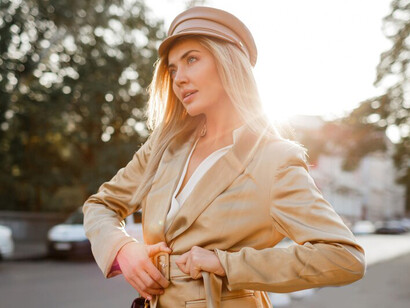 A stylish blonde woman in a beige cap and jacket strolls down the street, embracing an autumn look bathed in the warm glow of sunset, exuding timeless fashion