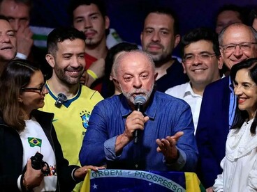 Lula discursa ao microfone, com uma das mãos estendida em um gesto de ênfase, enquanto aliados permanecem ao seu redor. À frente, uma bandeira brasileira cobre a parte superior da tribuna, trazendo à vista a inscrição “Ordem e Progresso”. A fotografia registra um momento de comunicação política em que a celebração da vitória se combina com a necessidade de apresentar os próximos passos. No horizonte de reflexão de Boaventura de Sousa Santos, a democracia não se esgota na escolha de representantes: depende também da capacidade de responder às desigualdades, ampliar a participação cidadã e submeter o exercício do poder ao controle público. A conquista eleitoral é, assim, um ponto de partida, não uma solução automática para os problemas do país
