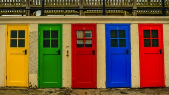 Uma sequência de portas de diferentes cores se alinha lado a lado em meio à paisagem urbana, criando uma composição marcada pela diversidade. Cada porta pode ser apenas uma escolha estética, mas também pode dizer algo sobre quem está do outro lado — afinal, como pergunta a autora, qual é a cor da porta da sua casa?