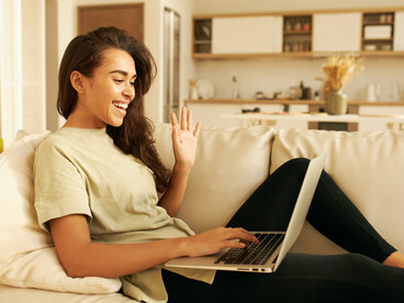 A young woman sits at her computer, waving at the screen during an AI therapy session focused on mental health