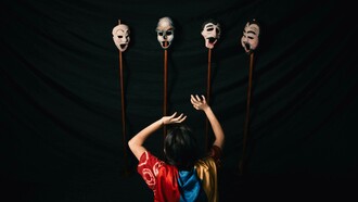 A woman in a robe performs with theater masks against a black background, embodying the tension between identity and performance