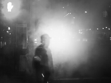 Una persona camina de noche entre el humo y las luces urbanas, parcialmente oculta por las sombras