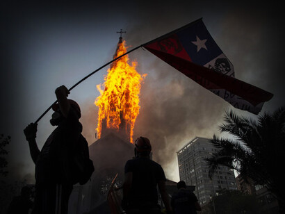 Hombre sujeta una bandera de Chile mientras arde en llamas una iglesia a sus espaldas
