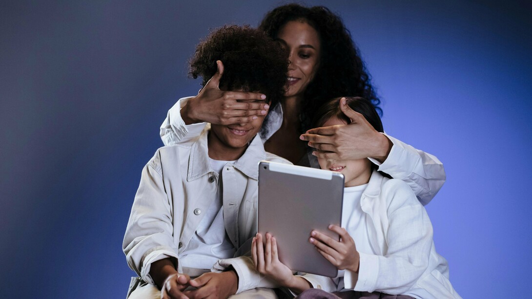 Uma mãe cobre os olhos dos filhos enquanto eles seguram um tablet diante da tela. Em tempos de hiperexposição digital, proteger também significa compreender o que os adolescentes consomem online. O gesto de proteção contrasta com a presença constante das telas no cotidiano infantil. A supervisão digital tornou-se parte essencial do cuidado emocional e cognitivo. Entre curiosidade e vulnerabilidade, crianças e adolescentes acessam conteúdos para os quais muitas vezes ainda não possuem maturidade emocional. O cérebro em formação responde intensamente aos estímulos digitais