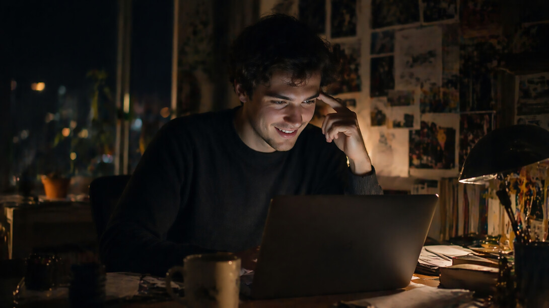 A young man sits at a dimly lit desk, smiling softly as he looks at his laptop, surrounded by scattered papers and warm ambient light