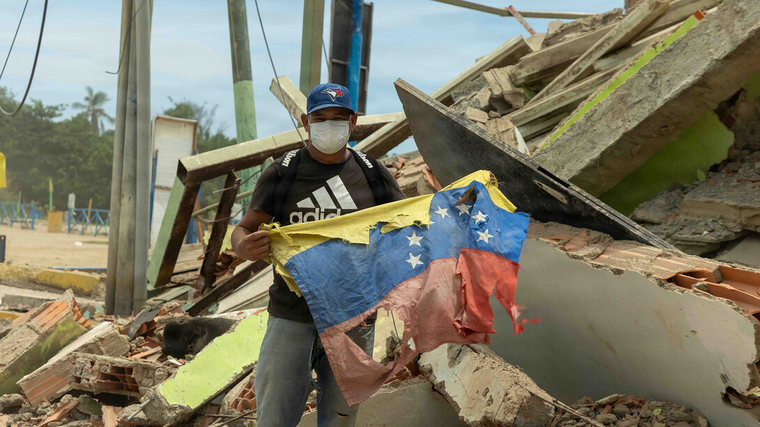 Ciudadano venezolano sostiene una bandera nacional junto a su restaurante dañado tras un terremoto en La Guaira, Venezuela, como signo de resiliencia. 28 de junio de 2026