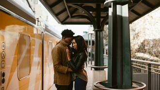 A couple hugs beside an orange train, sharing an emotional moment in a long-distance relationship