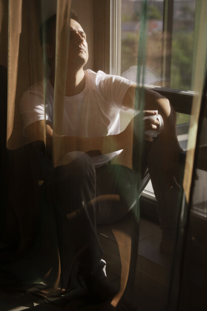 A man, deep in thought, sits by a window as gentle light and shadows from the curtains shape an emotional, introspective scene