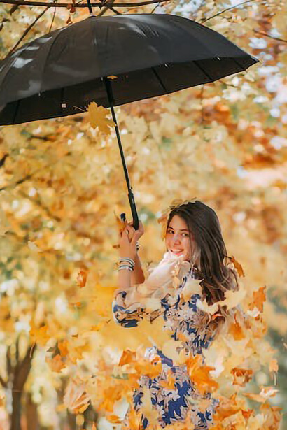 In this captivating photograph, a woman stands beneath the autumn rain, sheltered by an umbrella adorned with vibrant fall colors, creating a striking juxtaposition against the seasonal backdrop