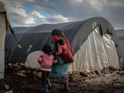 In Idlib, Idlib Governorate, Syria, a refugee camp with rows of tents sits in muddy conditions, with people gathered nearby navigating the difficult environment